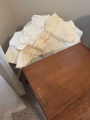 Collection of vintage white and cream baby garments, including hand-knit sweaters and cotton dresses, resting on a surface next to a...