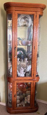 Tall oak corner display cabinet with curved glass side panels, glass shelving, and interior lighting, filled with various collectible...