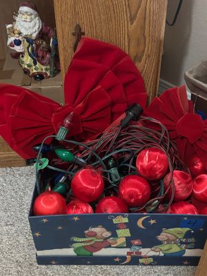 Box filled with Christmas decorations, including red velvet bows, red satin-wrapped ornaments, and strings of colorful outdoor lights.