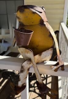 Whimsical metal frog garden statue, crafted from painted sheet metal with a weathered, rusted patina, designed to perch on a rail.