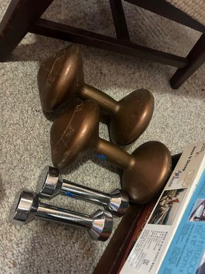 Two pairs of small hand weights, including bronze-colored coated dumbbells and chrome-plated steel dumbbells, resting on a carpeted floor...