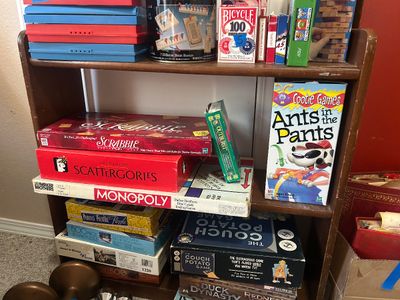 A vintage wooden shelf stacked with various classic board games, including Scrabble, Monopoly, Scattergories, and Ants in the Pants,...