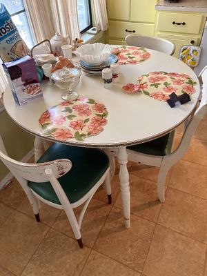 Vintage white round wooden dining table set with turned legs and dark green upholstered chairs, adorned with floral placemats and glassware.