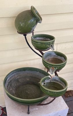 Tiered olive green ceramic water fountain on a metal stand, featuring a jug-shaped top spout draining into cascading bowls. Shows light...