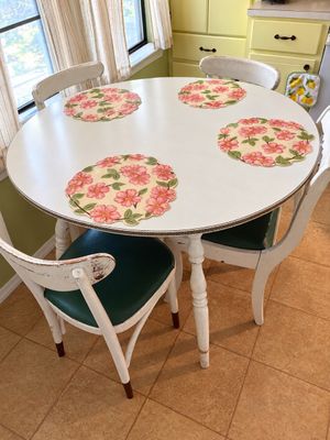 Vintage white round dining table paired with four distressed wooden chairs featuring green vinyl seats and four floral placemats.