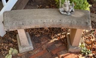 Weathered curved concrete garden bench with carved detail along the front edge, topped with a small frog statue.