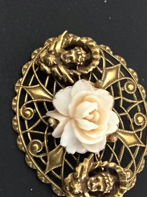 Sale Photo Thumbnail #44: An intricate oval vintage brooch featuring detailed gold-tone filigree metalwork, a central carved faux-coral or resin rose bloom, and two cherub/putti figures on either side. Features a rollover safety clasp on the reverse. Shows minor signs of vintage w