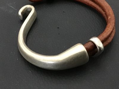 Sale Photo Thumbnail #183: A stylish bracelet featuring a thick, curved silver-tone metal hook clasp secured to a double strand of brown leather cord. Includes a silver-tone accent slider with a dangling bezel-set round tiger's eye or brown stone charm. Clasp hook measures approxim