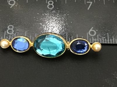 Sale Photo Thumbnail #243: A vintage gold tone bar brooch featuring three oval-cut blue glass rhinestones in varying shades of aqua, light blue, and deep sapphire blue, flanked by two small faux pearls at either end. The pin features a textured back with a standard safety bar clasp