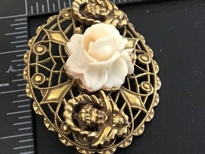 Sale Photo Thumbnail #42: An intricate oval vintage brooch featuring detailed gold-tone filigree metalwork, a central carved faux-coral or resin rose bloom, and two cherub/putti figures on either side. Features a rollover safety clasp on the reverse. Shows minor signs of vintage w