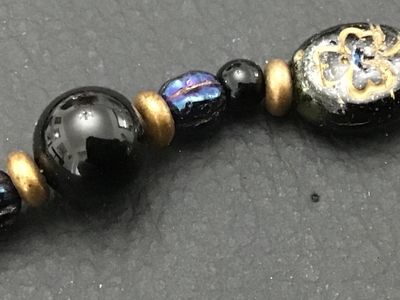 Sale Photo Thumbnail #249: Beaded strand bracelet featuring oval black beads decorated with gold-tone shamrock motifs, iridescent purple/blue accent beads, smooth black spacer beads, and brass-tone metal spacers. Fitted with a lobster claw clasp marked 'GF' (gold-filled). Measures 