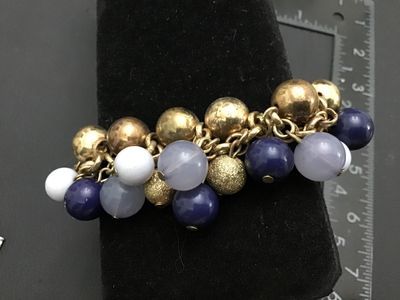 Sale Photo Thumbnail #270: A chunky gold-tone link bracelet featuring an array of dangling bead charms including textured gold-tone metal spheres, smooth dark blue beads, translucent greyish-blue beads, and white beads. The bracelet measures approximately 7 inches in circumference.