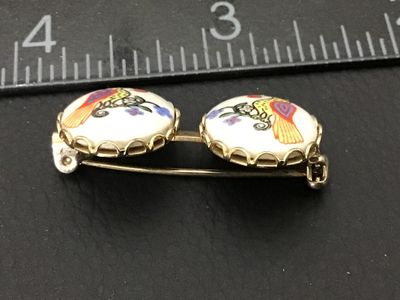 Sale Photo Thumbnail #199: A vintage double-oval brooch featuring hand-painted or transfer porcelain cabochons set in gold-tone metal. Each oval displays a colorful bird perched on a floral branch. Measures approximately 1.25 inches across. The pin back and C-clasp are in functiona