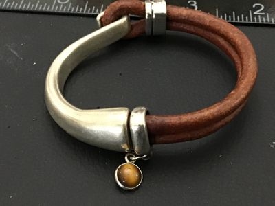 Sale Photo Thumbnail #181: A stylish bracelet featuring a thick, curved silver-tone metal hook clasp secured to a double strand of brown leather cord. Includes a silver-tone accent slider with a dangling bezel-set round tiger's eye or brown stone charm. Clasp hook measures approxim