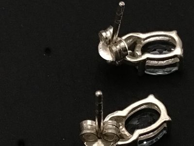 Sale Photo Thumbnail #190: Pair of  silver stud earrings featuring oval-cut light blue gemstones, likely aquamarine or blue topaz, set in four-prong settings with small decorative accents at the top. Includes matching silver friction butterfly backs. Good pre-owned condition with m