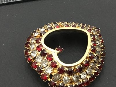 Sale Photo Thumbnail #163: A vintage gold-tone heart-shaped brooch pin encrusted with red and clear/smokey rhinestones set in prong settings. Features a rollover safety clasp on the back. Measures approximately 1.5 inches wide by 1.5 inches tall. Overall good vintage condition with