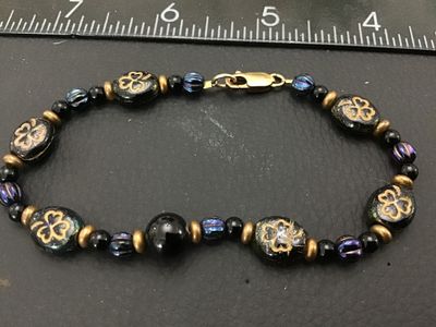 Sale Photo Thumbnail #247: Beaded strand bracelet featuring oval black beads decorated with gold-tone shamrock motifs, iridescent purple/blue accent beads, smooth black spacer beads, and brass-tone metal spacers. Fitted with a lobster claw clasp marked 'GF' (gold-filled). Measures 