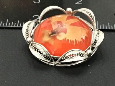 Sale Photo Thumbnail #156: A vintage silver-tone filigree brooch featuring a center domed cabochon with an embedded orange botanical/flower design. The back features a standard pin bar mechanism along with a small bail, allowing it to be worn as either a brooch or a pendant. Measur