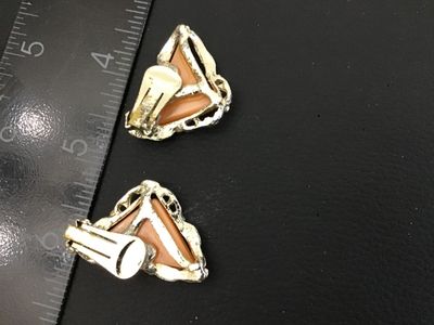 Sale Photo Thumbnail #256: A pair of vintage gold-tone clip-on earrings featuring triangular peach-pink cabochons or resin centers set in a textured, scroll-style border. Each earring measures approximately 1 inch in length. The clip backs show light vintage wear and finish loss on