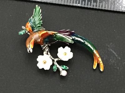 Sale Photo Thumbnail #232: Colorful enameled costume jewelry brooch featuring a long-tailed bird perched on a flowering branch. Styled with vibrant green, orange, and purple enamel, white carved flower accents, and green/clear rhinestones on silver-tone metal backing. Features a st