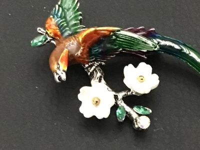 Sale Photo Thumbnail #234: Colorful enameled costume jewelry brooch featuring a long-tailed bird perched on a flowering branch. Styled with vibrant green, orange, and purple enamel, white carved flower accents, and green/clear rhinestones on silver-tone metal backing. Features a st