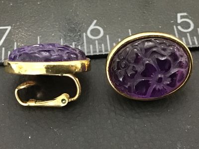 Sale Photo Thumbnail #165: Pair of vintage gold-tone oval clip-on earrings featuring carved purple glass inserts with floral patterns. Each earring measures approximately 0.75 inches long. Backs feature a textured sunburst finish and clip mechanisms. Light vintage wear to metal fin