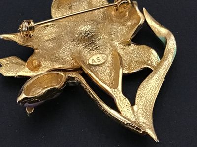 Sale Photo Thumbnail #126: A vintage gold-tone costume jewelry brooch featuring a pink and white enamel lily flower with a green enameled leaf stem and a green rhinestone-accented dragonfly resting on the petals. The back is signed 'AS ©' and features a secure pin stem and rollove