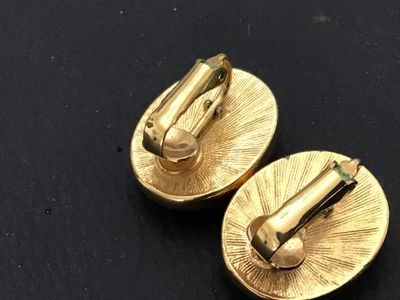 Sale Photo Thumbnail #166: Pair of vintage gold-tone oval clip-on earrings featuring carved purple glass inserts with floral patterns. Each earring measures approximately 0.75 inches long. Backs feature a textured sunburst finish and clip mechanisms. Light vintage wear to metal fin
