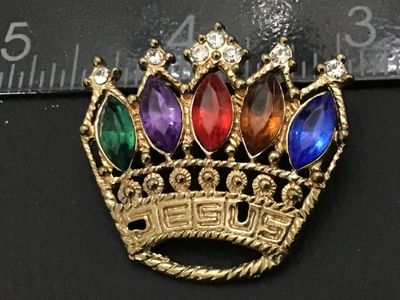 Sale Photo Thumbnail #88: Gold-tone metal brooch featuring a crown design with 'JESUS' spelled along the band. Set with multi-colored navette rhinestones in green, purple, red, topaz, and blue, topped with clear accent rhinestones. Measures approximately 1.5 inches wide. Standard 