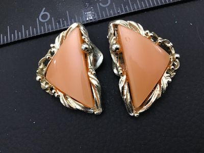 Sale Photo Thumbnail #253: A pair of vintage gold-tone clip-on earrings featuring triangular peach-pink cabochons or resin centers set in a textured, scroll-style border. Each earring measures approximately 1 inch in length. The clip backs show light vintage wear and finish loss on