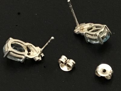 Sale Photo Thumbnail #191: Pair of  silver stud earrings featuring oval-cut light blue gemstones, likely aquamarine or blue topaz, set in four-prong settings with small decorative accents at the top. Includes matching silver friction butterfly backs. Good pre-owned condition with m