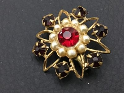 Sale Photo Thumbnail #285: Vintage gold-tone brooch featuring a central bright red rhinestone framed by faux pearls, surrounded by dark red/garnet-colored rhinestone accents in a starburst or flower motif. Shows slight vintage wear and patina to the gold-tone metal on the back. Fea