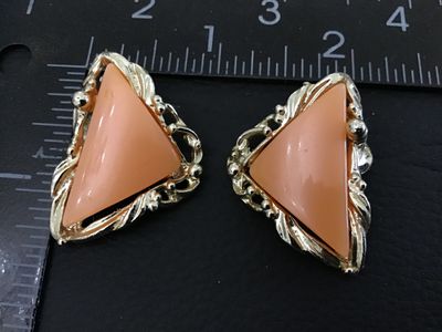 Sale Photo Thumbnail #254: A pair of vintage gold-tone clip-on earrings featuring triangular peach-pink cabochons or resin centers set in a textured, scroll-style border. Each earring measures approximately 1 inch in length. The clip backs show light vintage wear and finish loss on