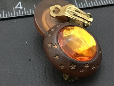 Sale Photo Thumbnail #284: Pair of vintage round clip-on earrings featuring large faceted amber-colored glass or lucite center stones set in dark brown wooden frames detailed with carved star motifs. Equipped with gold-tone clip-on backs. Each earring measures approximately 1.25 in