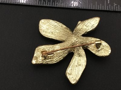 Sale Photo Thumbnail #98: A floral brooch featuring layered petals in light purple and white enamel with gold-tone edging. The back shows a textured gold-tone finish with a standard pin stem and rollover safety clasp. Measures approximately 2.25 inches across. Overall good pre-own