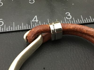 Sale Photo Thumbnail #182: A stylish bracelet featuring a thick, curved silver-tone metal hook clasp secured to a double strand of brown leather cord. Includes a silver-tone accent slider with a dangling bezel-set round tiger's eye or brown stone charm. Clasp hook measures approxim