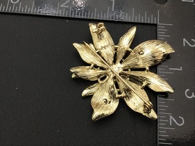 Sale Photo Thumbnail #155: A festive vintage gold-tone brooch shaped like a poinsettia flower featuring red and green enamel layered petals. The center is detailed with a cluster of small sparkling clear rhinestones. Features a standard pin back clasp mechanism. Measures approximat