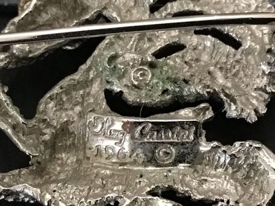 Sale Photo Thumbnail #245: Vintage Oleg Cassini dragon or mythical creature brooch pin, dated 1964 on back plaque. Textured silver-tone finish accenting blue, light blue, and red rhinestones. Measures approximately 1.75 inches across. Good vintage condition with working pin mechani