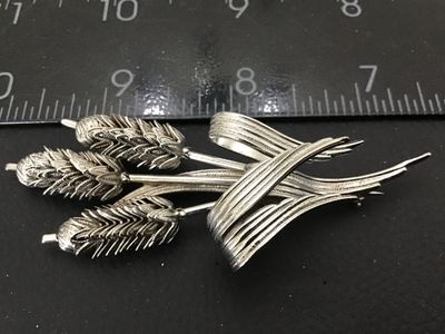 Sale Photo Thumbnail #58: A silver-tone costume jewelry brooch designed as a bundle of three wheat stalks tied with a textured ribbon motif. Features detailed textured grains and a pin-back closure on the reverse. Measures approximately 3 inches in length. In good condition with m