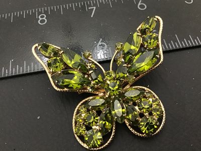 Sale Photo Thumbnail #24: A vintage gold-tone butterfly brooch featuring multi-faceted olive/peridot green rhinestones in marquise and round cuts. The wings have a textured rope-style border. Measures approximately 2.5 inches wide by 2 inches tall. Features a standard rollover saf