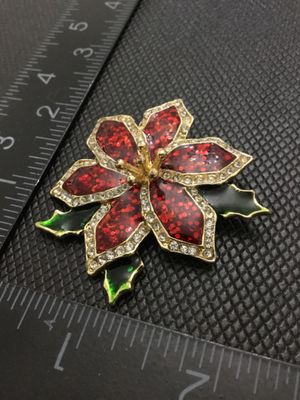 Sale Photo Thumbnail #209: A festive gold-tone holiday poinsettia brooch featuring red glitter enamel petals bordered by clear rhinestones and green enamel holly leaf accents. Marked '©TC' on the reverse side. Has a standard pin back with standard clasp function. Measures approxim