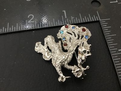Sale Photo Thumbnail #247: Vintage Oleg Cassini dragon or mythical creature brooch pin, dated 1964 on back plaque. Textured silver-tone finish accenting blue, light blue, and red rhinestones. Measures approximately 1.75 inches across. Good vintage condition with working pin mechani