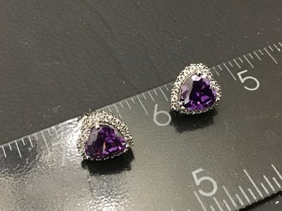 Sale Photo Thumbnail #107: Pair of silver-tone stud earrings featuring heart-cut purple central stones surrounded by a halo of clear accent stones. Secured with friction post backs. Each earring measures approximately 1/2 inch in size. Good condition with light wear.