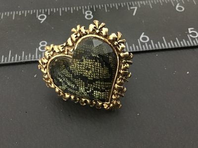 Sale Photo Thumbnail #8: A signed Betsey Johnson costume jewelry ring featuring a large heart-shaped focal piece with a faceted cover and dark pattern underneath, surrounded by a gold-tone ornate bow and heart border. The inside band is stamped 'Betsey Johnson' and set with a sig