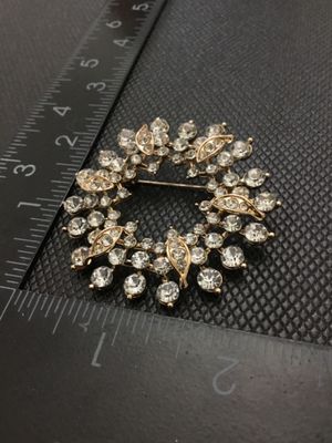 Sale Photo Thumbnail #219: A vintage gold-tone circular wreath brooch or pin, encrusted with round clear rhinestones and featuring leaf-like textured accents with embedded stones. Measures approximately 2 inches in diameter. Features a rollover safety clasp on the back. Appears to 