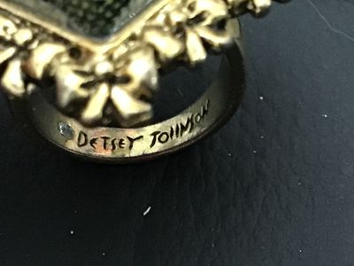Sale Photo Thumbnail #11: A signed Betsey Johnson costume jewelry ring featuring a large heart-shaped focal piece with a faceted cover and dark pattern underneath, surrounded by a gold-tone ornate bow and heart border. The inside band is stamped 'Betsey Johnson' and set with a sig