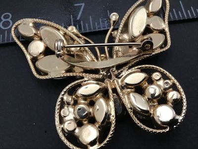 Sale Photo Thumbnail #26: A vintage gold-tone butterfly brooch featuring multi-faceted olive/peridot green rhinestones in marquise and round cuts. The wings have a textured rope-style border. Measures approximately 2.5 inches wide by 2 inches tall. Features a standard rollover saf
