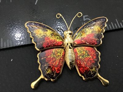 Sale Photo Thumbnail #19: Gold-tone metal butterfly brooch featuring colorful red, orange, and gold glitter inlay or enamel fill on the wings. Measures approximately 2 inches wide by 2 inches tall. Features a standard rollover safety pin closure on the back. Good pre-owned conditi