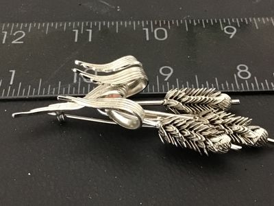 Sale Photo Thumbnail #60: A silver-tone costume jewelry brooch designed as a bundle of three wheat stalks tied with a textured ribbon motif. Features detailed textured grains and a pin-back closure on the reverse. Measures approximately 3 inches in length. In good condition with m