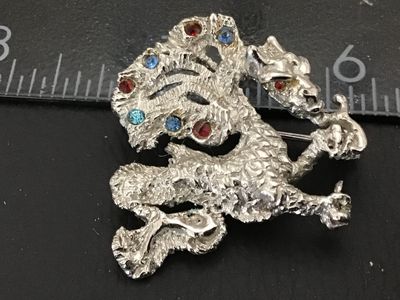 Sale Photo Thumbnail #244: Vintage Oleg Cassini dragon or mythical creature brooch pin, dated 1964 on back plaque. Textured silver-tone finish accenting blue, light blue, and red rhinestones. Measures approximately 1.75 inches across. Good vintage condition with working pin mechani